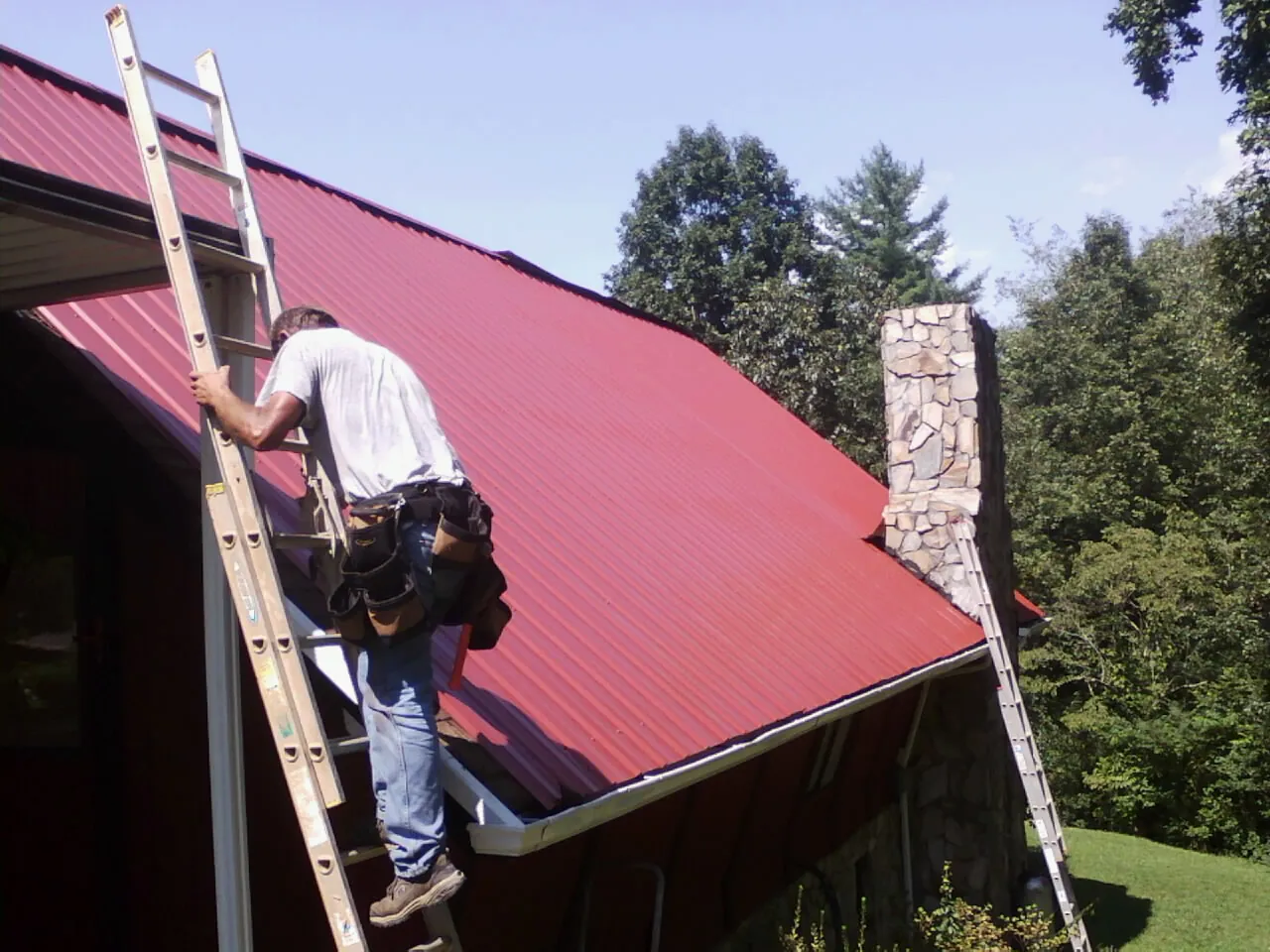 Roofer on a ladder beside a red metal roof and stone chimney — Soffit & Fascia Repair by Roofing Brothers in Saxton